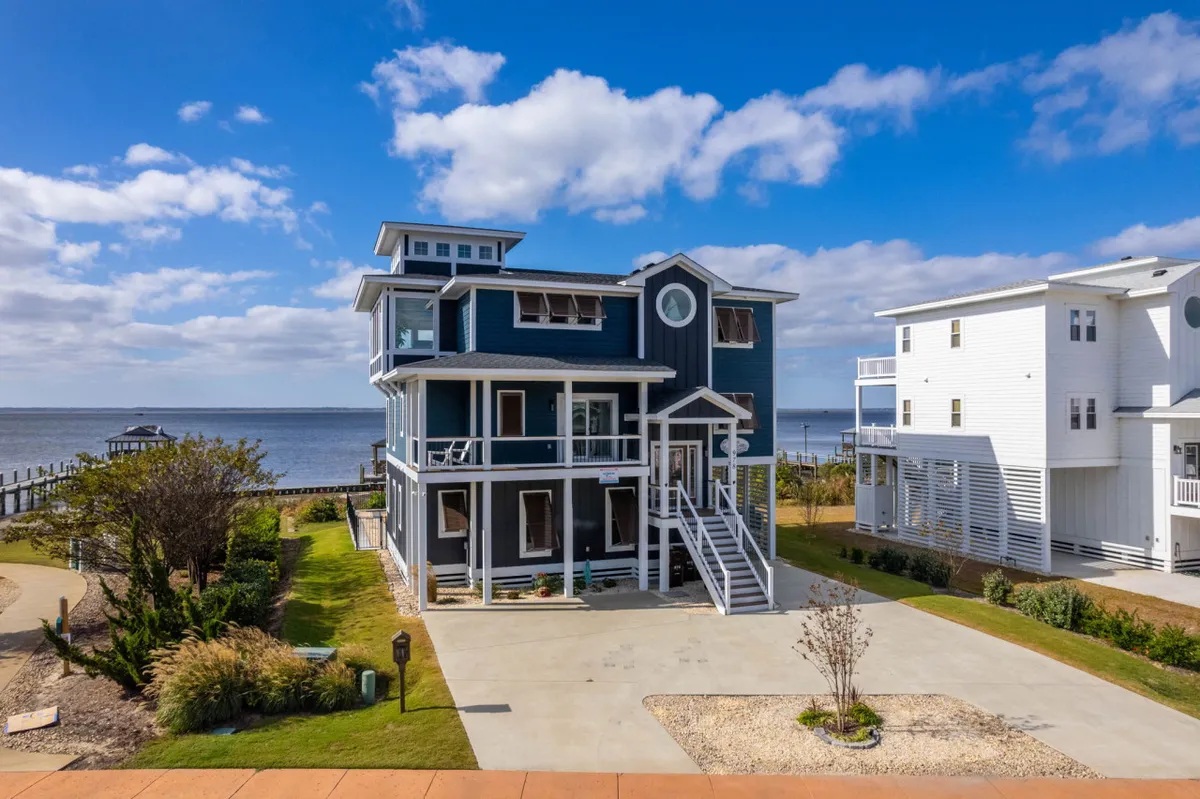This image showcases the blue facade of the house, a spacious porch, and a view of the water. The porch is set up for relaxation and gatherings, with ample outdoor space for family, friends, and pets to enjoy the fresh air just steps from the beach.