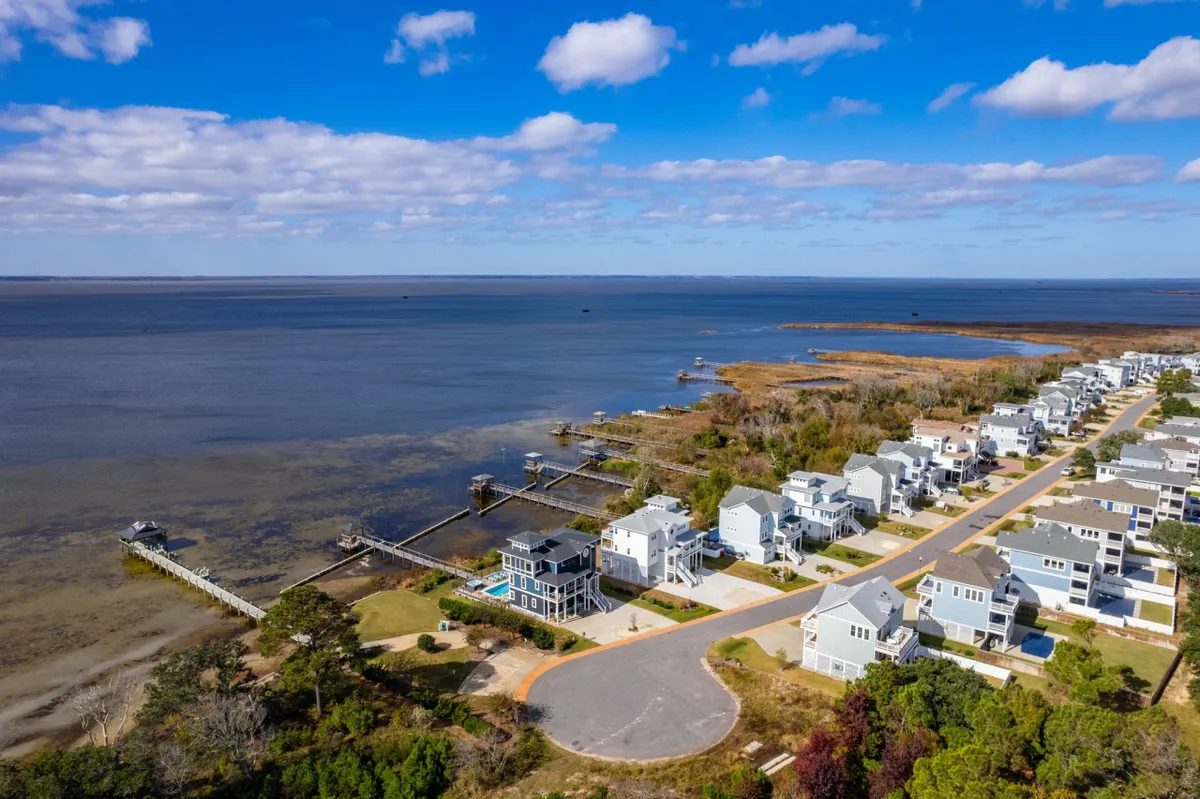 A scenic exterior view featuring waterfront homes, marshland, a pier, and a road leading to the coast. The tranquil setting highlights the area’s dual waterfront and beach access, with open skies and space for exploration and pet-friendly walks.