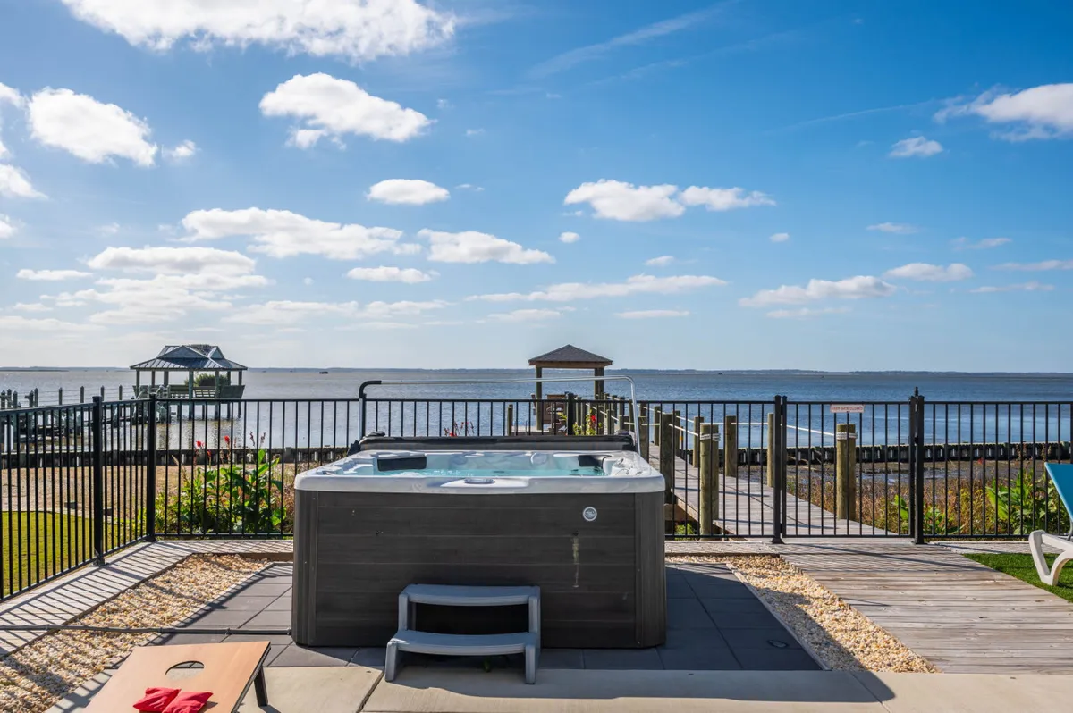 The photo captures a tranquil deck with a bubbling hot tub overlooking the water, surrounded by a fence for privacy. The scene suggests a peaceful soak with beautiful views, ideal for year-round relaxation.