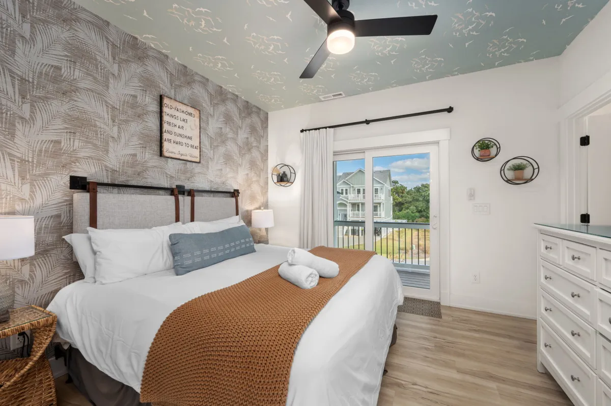This image captures a serene king bedroom featuring a bed with decorative pillows, a woven throw blanket, and a ceiling with a floral pattern. Large sliding glass doors fill the room with natural light and offer picturesque views, reinforcing the sense of tranquility and openness.