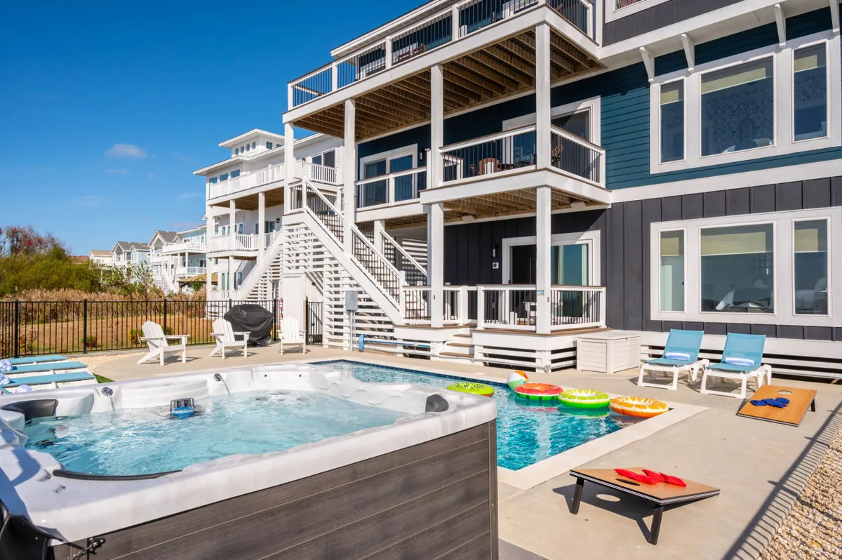 The hero image showcases a spacious backyard with a sparkling swimming pool and inviting hot tub, surrounded by colorful pool floats and comfortable deck chairs. The pool area is the heart of the outdoor space, set against a backdrop of the upper deck where sunset views over Currituck Sound are visible. The mood is playful yet relaxing, suggesting opportunities for swimming, lounging, and enjoying poolside barbecues or stargazing in the evening.