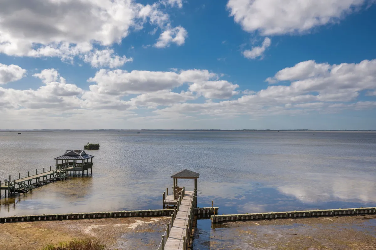A tranquil dock extends over the calm waters of Currituck Sound, with a pavilion at the end and the water reflecting the sky and clouds above. The setting is peaceful and inviting, suggesting opportunities for fishing, yoga, or simply soaking in the views.