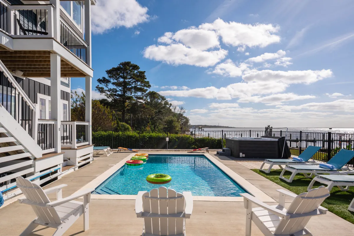 A sunlit outdoor pool area with lounge chairs arranged around the water, set against a backdrop of sparkling blue water views. The relaxed setup invites guests to swim, sunbathe, or unwind by the pool while enjoying the tranquil coastal scenery.