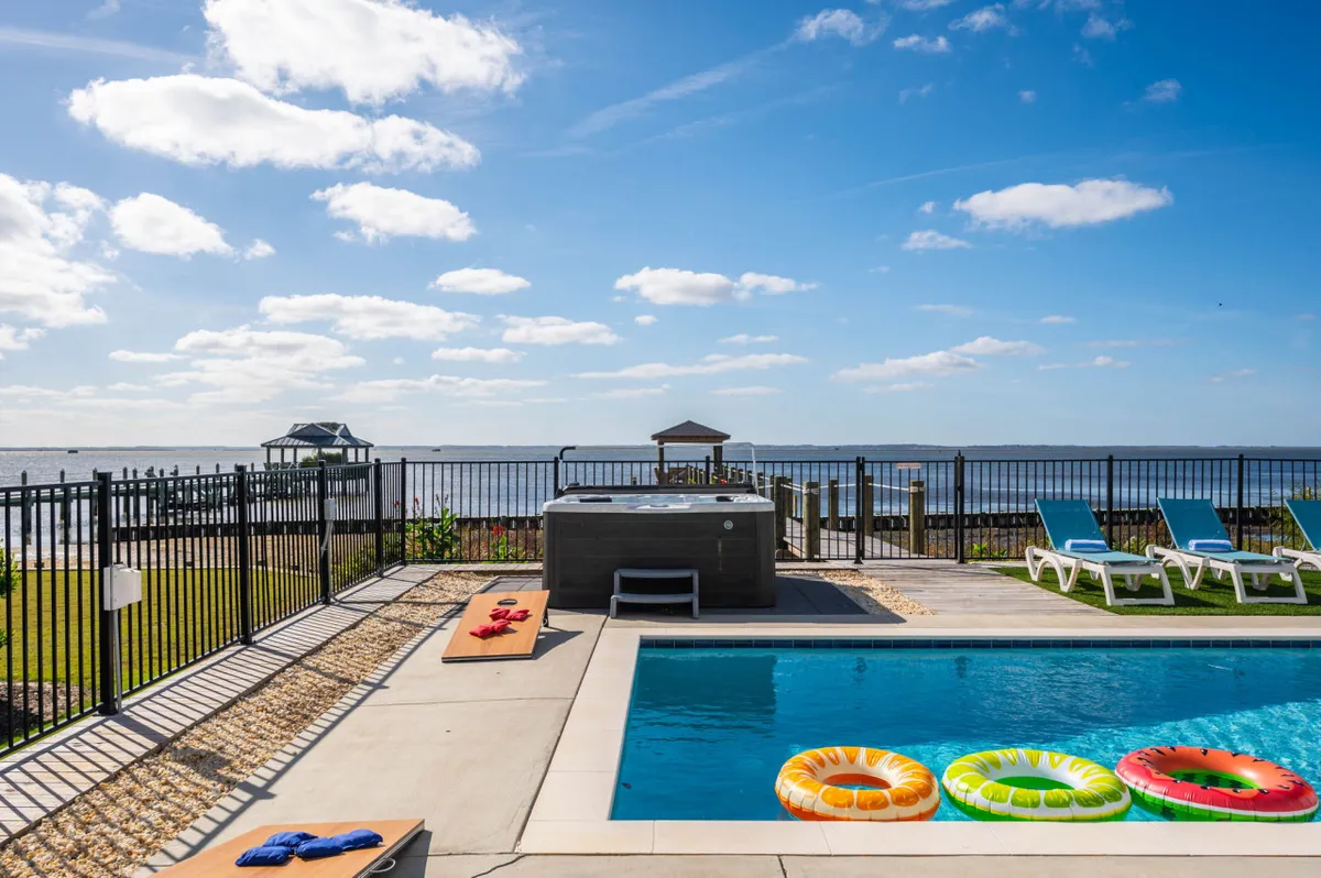 An inviting exterior scene with a sparkling pool, a bubbling hot tub, colorful floaties, lounge chairs, and a sweeping view of the waterfront landscape. Sunset colors hint at evening relaxation.