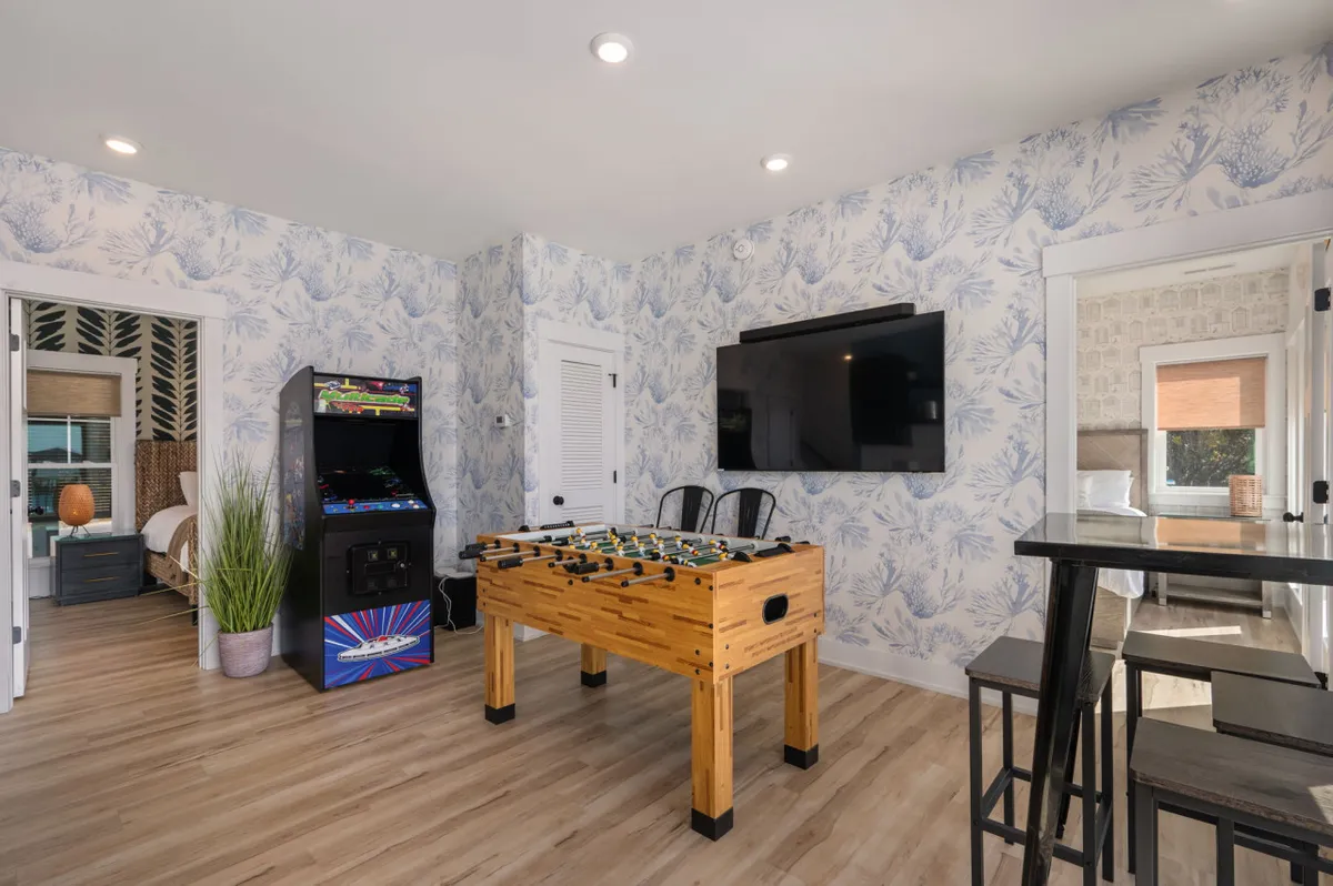 A vibrant game room featuring a foosball table, an arcade game machine, a large flat-screen TV, and decorative wallpaper. The space is well-lit and arranged for both active play and socializing.