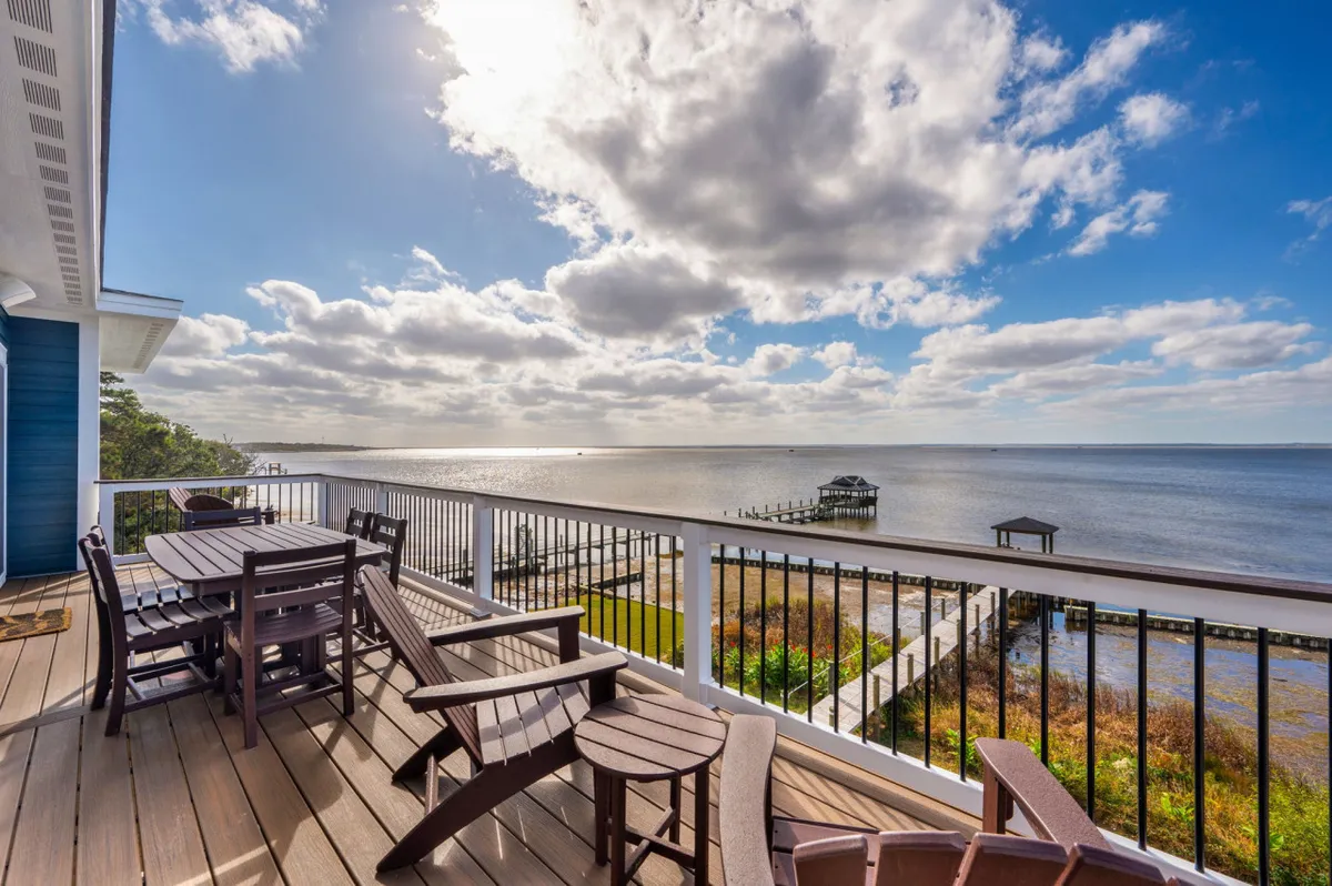 A waterfront deck furnished with seating and a table, offering sweeping views of the calm water and a distant dock. The scene suggests opportunities for relaxing, sharing meals outside, and soaking up sunsets after a day at the beach.