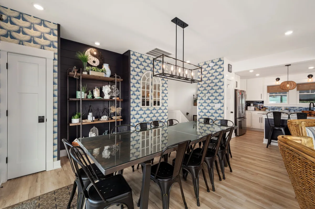 A spacious dining area featuring a large table set beneath a striking chandelier, complemented by textured wallpaper and a decorative wall shelf. The setup suggests ample space for group meals, festivities, and lingering conversations with water views suggested by the light-filled backdrop.
