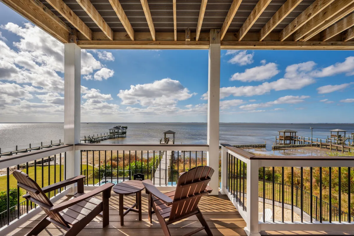 A peaceful deck scene with wooden chairs overlooks serene water and a deck railing, suggesting a tranquil atmosphere for relaxing while taking in the view, especially at sunset. The setting feels intimate and accessible, ideal for unwinding after a walk or beach play with pets.
