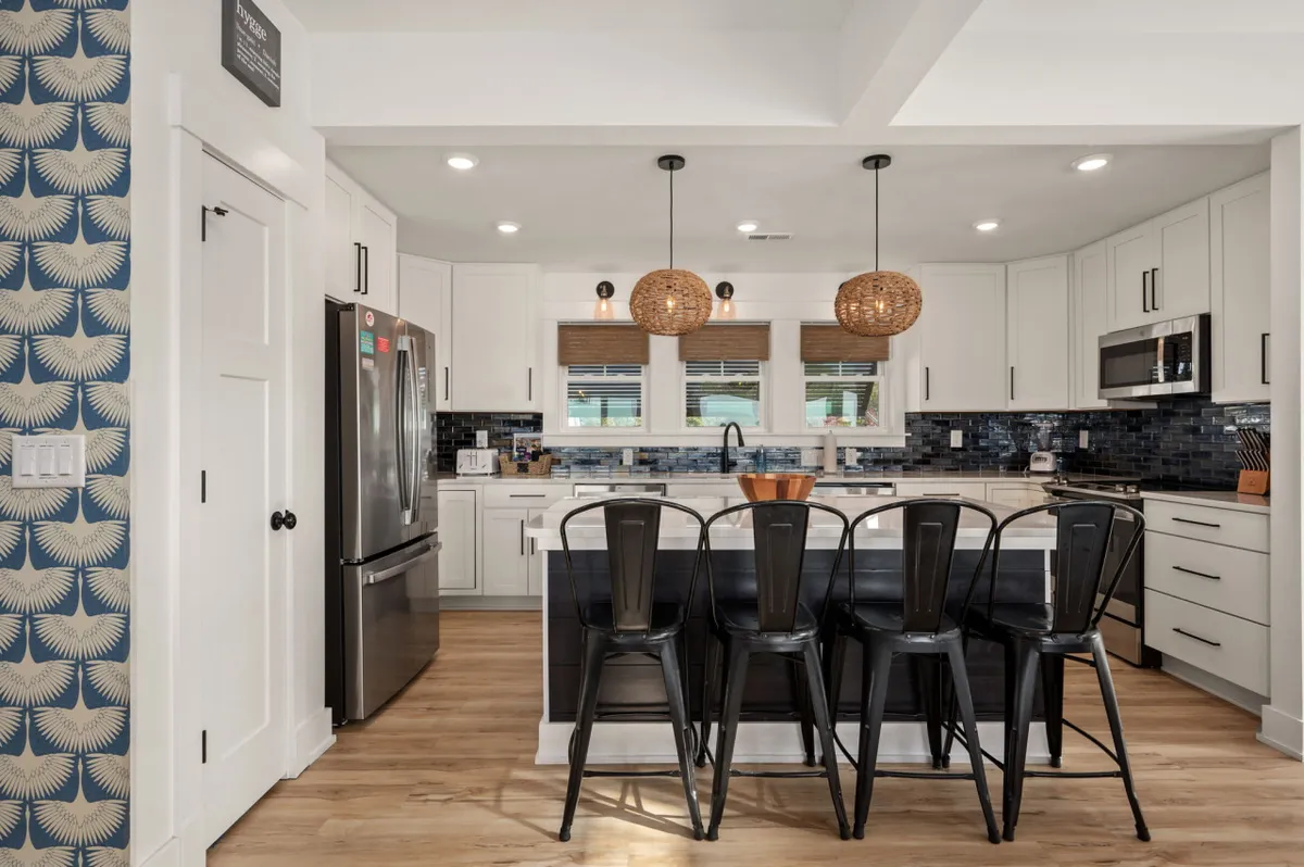 A vibrant, welcoming kitchen with a spacious island at the center, lined with black metal bar stools and illuminated by woven pendant lights. The kitchen features a modern blue tile backsplash and is equipped with stainless steel appliances, creating a perfect space for group cooking and casual gatherings.