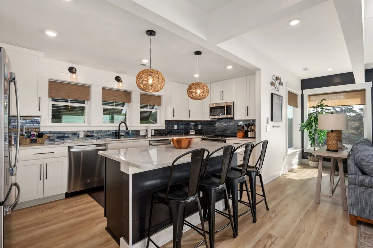 A wide view of a beautifully designed kitchen with a central island counter and bar seating, pendant light fixtures, and modern stainless steel appliances. The blue-tiled backsplash ties in a coastal vibe, while the layout invites both cooking and socializing.
