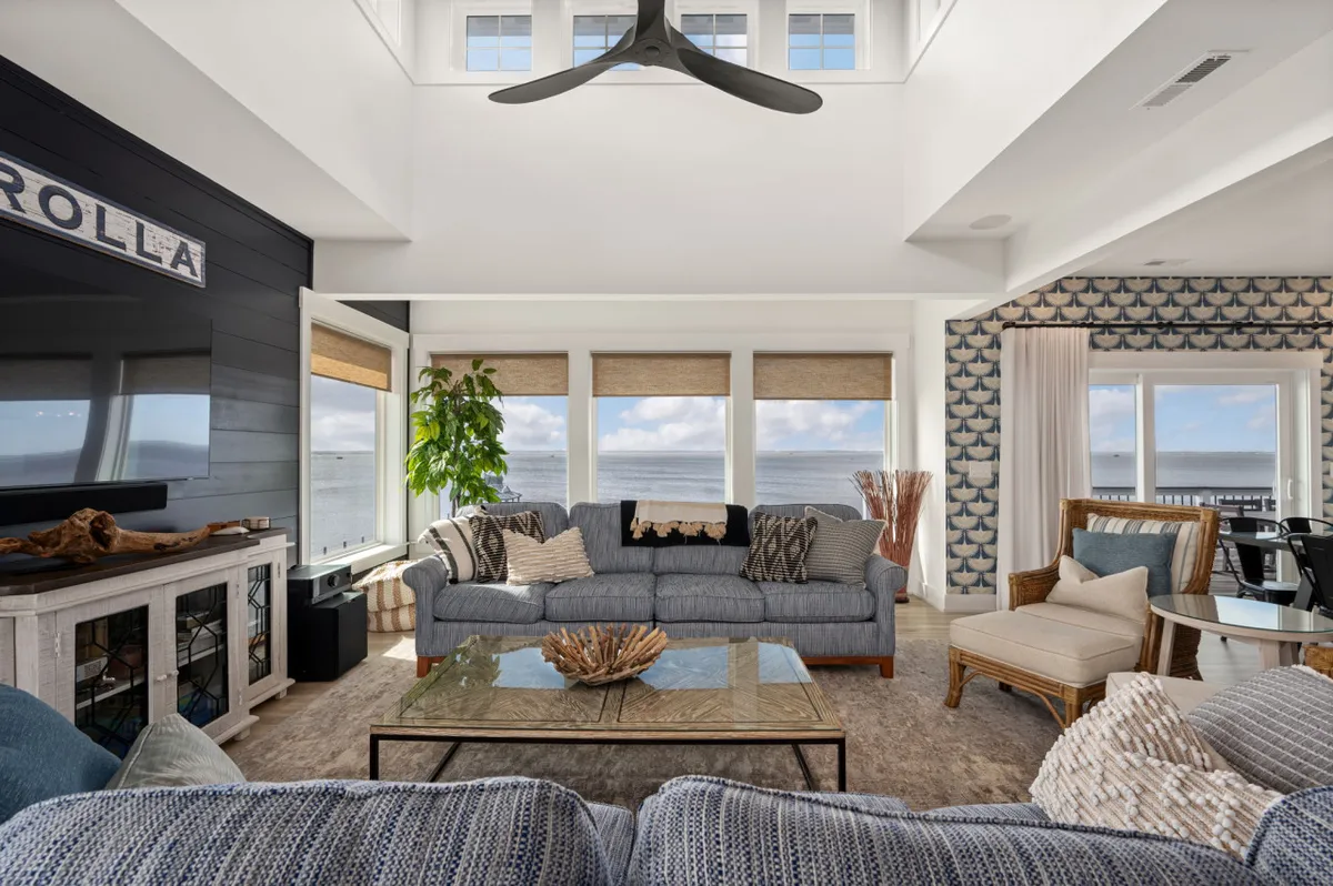 A casually elegant living area with a comfortable blue sofa, glass coffee table, decorative fish-patterned wall, indoor plants, and large windows revealing an ocean view. The space feels welcoming and ideal for both relaxation and entertainment.