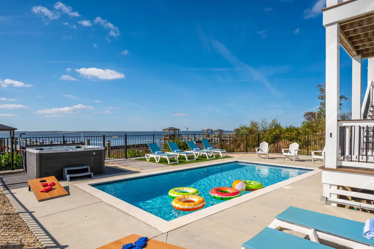 This exterior image displays a private swimming pool and hot tub surrounded by lounge chairs and colorful pool floats, with a serene water view in the background. The setting is vibrant and inviting, ideal for relaxation and recreation.