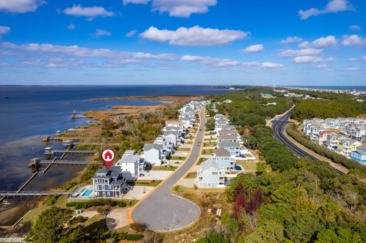 This image captures a wide view of the waterfront, including the neighborhood layout with houses, a road, marshland, and calm water. The presence of open grassy areas and a gentle shoreline evokes the feel of an inviting, natural environment that is ideal for walking, outdoor play, and enjoying the water's edge.