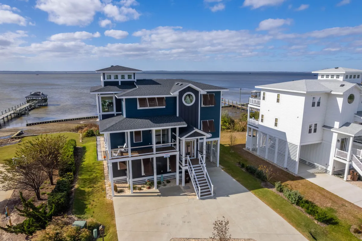 The home sits directly on the waterfront, with a spacious porch overlooking the sound and a dock stretching into the water. The scene captures the house, dock, and surrounding calm waters, perfect for relaxing or gathering with loved ones.