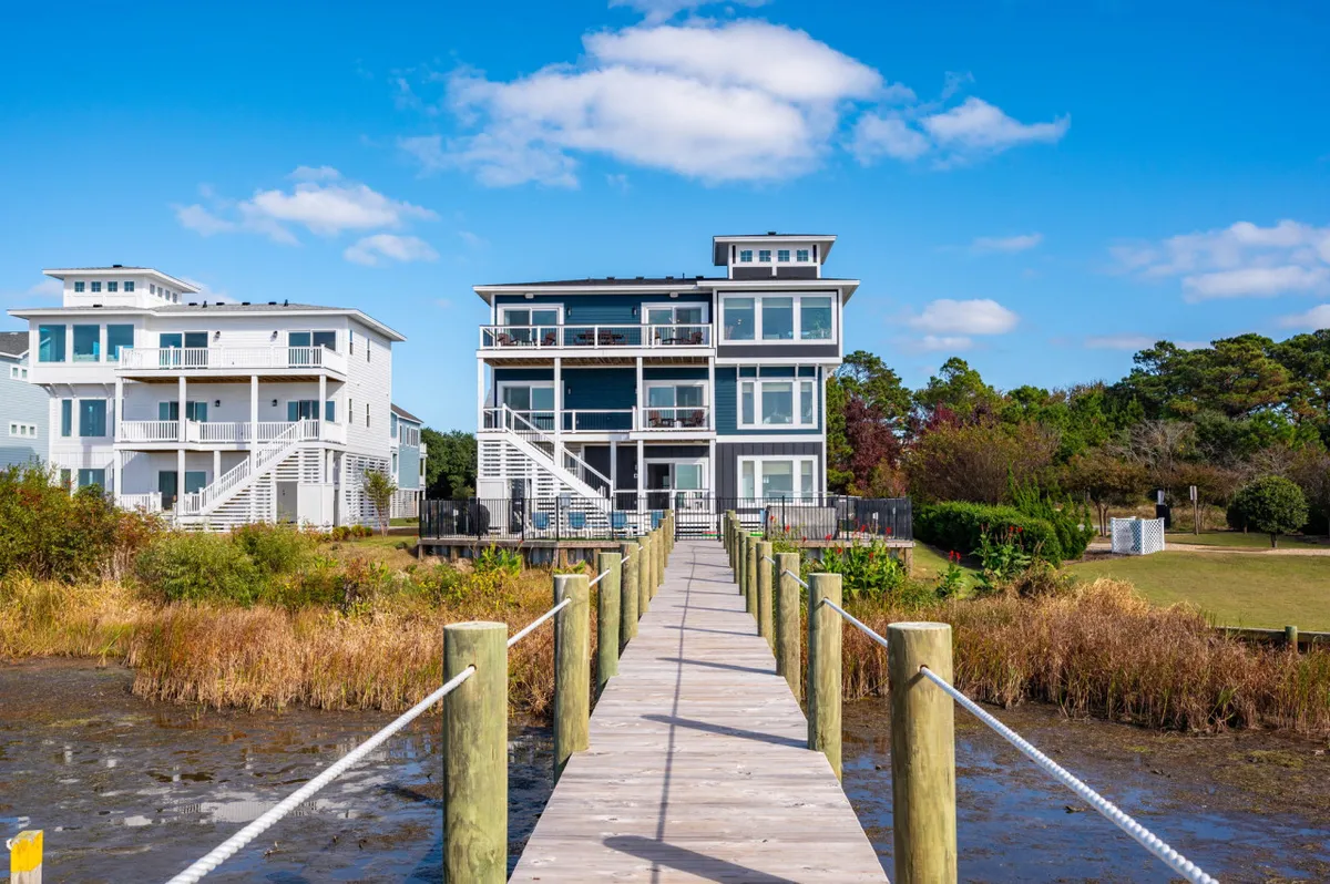 The exterior of a spacious home with multiple balconies, surrounded by lush natural vegetation. A wooden walkway leads to the water, inviting guests to enjoy fishing, sunset views, and gatherings by the shore.
