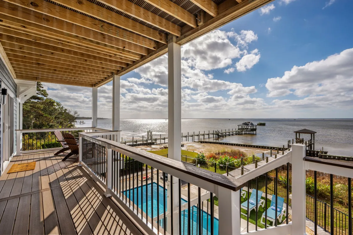 A wooden deck with a pool overlooking the water, a fishing pier visible in the distance, and comfortable seating arranged for relaxation. The tranquil setting showcases the seamless blend of outdoor leisure and scenic waterfront living.